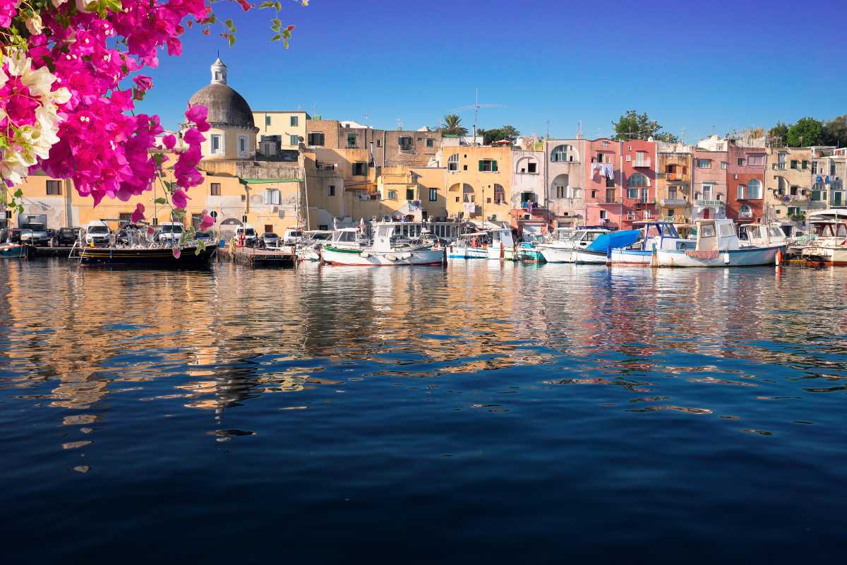 Bambini che partecipano a un laboratorio creativo all’aperto a Procida, tavolo con materiali artistici, colori e sorrisi, case colorate sullo sfondo
